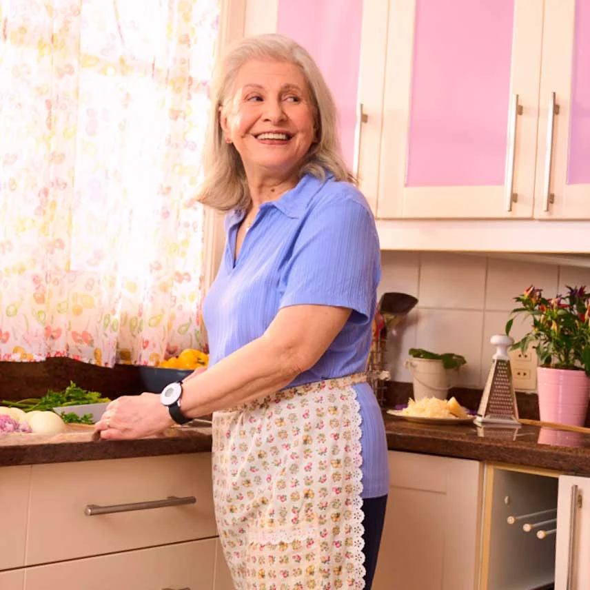 Dona Helena sorrindo na cozinha enquanto segue sua rotina.